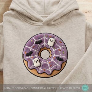 Op de afbeelding: Een beige hoodie met een grote geborduurde afbeelding op de borst van een paarse donut met een wit spinnenwebpatroon, kleurrijke hagelslag, twee kleine witte spookfiguren en drie zwarte silhouetten van vleermuizen in het midden.