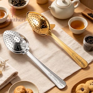 May include: Two metal tea infuser spoons, one gold-toned and one silver-toned, featuring perforated oval bowls and long handles. The silver handle is engraved with Tea Makes Home, while the gold handle is engraved with Tea With You.