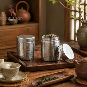 May include: Two cylindrical silver-toned metal canisters with textured surfaces. One canister is closed and features the name William engraved on the side. The other canister is open, filled with loose green tea leaves, with its lid showing the name Benjamin.