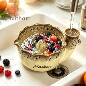 May include: A cream-colored ceramic berry colander with a brown speckled rim and a built-in water funnel. The bowl features the name Madison in black script and contains water with fresh blackberries, raspberries, and blueberries inside.