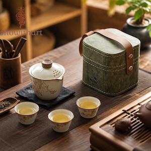Könnte beinhalten: Ein Keramik-Teeservice mit einem Gaiwan mit Deckel und drei kleinen Tassen, alle mit zarten rosa Blumenmustern verziert. Das Set enthält eine strukturierte olivgrüne Tragetasche mit braunem Lederriemen und dem Schriftzug Natural Chai.