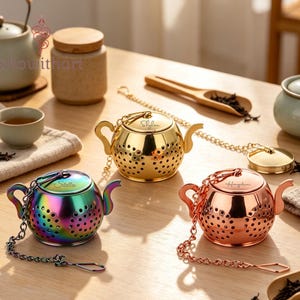 Könnte beinhalten: Drei teekannenförmige Tee-Eier mit Ketten und Deckeln. Eines ist goldfarben mit dem Namen Skylar, eines ist regenbogenfarben schillernd mit dem Namen Stella und eines ist roségold mit dem Namen Hayden. Jedes Tee-Ei hat einen perforierten Körper.