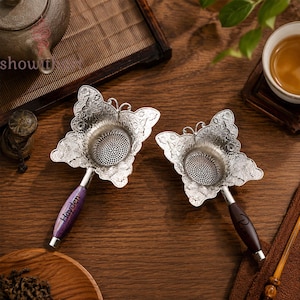 May include: Two silver-toned metal tea strainers shaped like butterflies with intricate floral embossing. One strainer features a purple marbled handle with the name Hayden printed on it, while the other has a dark brown wooden handle.