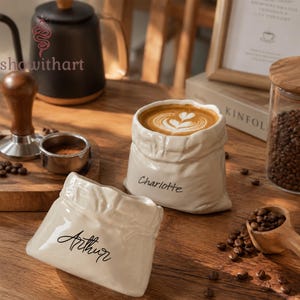 May include: Two off-white ceramic mugs shaped like crumpled fabric sacks. One mug is filled with coffee featuring latte art and has the name Charlotte printed on the side. The other empty mug has the name Arthur printed on the side in black script.