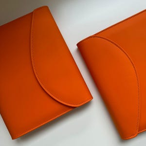 Può includere: Due portadocumenti in pelle arancione brillante con chiusure a patta curva. Le cartelle sono esposte su una superficie bianca, mostrando la loro consistenza liscia e i bordi cuciti. Il design è minimalista e moderno.