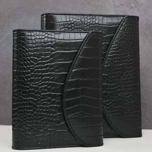 Op de afbeelding: Twee zwarte leren organizers met een krokodillenleertextuur. De organizers zijn rechthoekig met gebogen klepsluitingen. De grotere organizer staat achter de kleinere, beide op een wit oppervlak.
