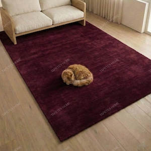 May include: A burgundy rectangular rug with a sleeping orange cat. A light-colored two-seater sofa with a wooden frame sits above the rug. The rug is on a wooden floor, and a window with white curtains is visible.