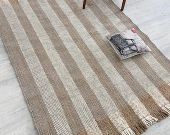 May include: A rectangular rug with vertical stripes in shades of brown and cream. The rug has a fringed edge and is placed on a light-colored floor. A wooden chair with a black seat and a decorative pillow are on the rug.