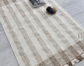 May include: A rectangular, striped rug with a cream and brown color scheme. The rug features vertical cream stripes and brown stripes, with a fringed edge. A small pillow and a basket of yarn balls are on the rug.