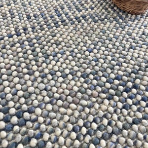 May include: A textured rug featuring a pattern of small, round wool balls in shades of blue, grey, and white. A small basket with blue and beige yarn balls sits in the upper right corner. The rug has a dense, tactile appearance.