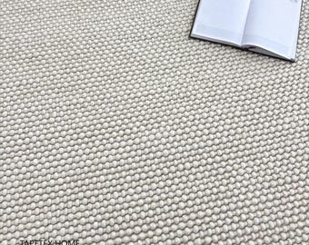 Puede incluir: Una alfombra texturizada de color blanquecino con una superficie densa y nudosa. Un cuaderno abierto con una cubierta negra descansa sobre la alfombra, mostrando páginas en blanco con líneas. La textura de la alfombra crea una sensación acogedora e invitadora.