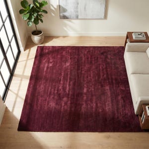 May include: A large, rectangular burgundy rug dominates the room's center. The rug has a soft, textured appearance and is placed on a light-colored hardwood floor. A neutral-toned sofa and side table are partially visible.