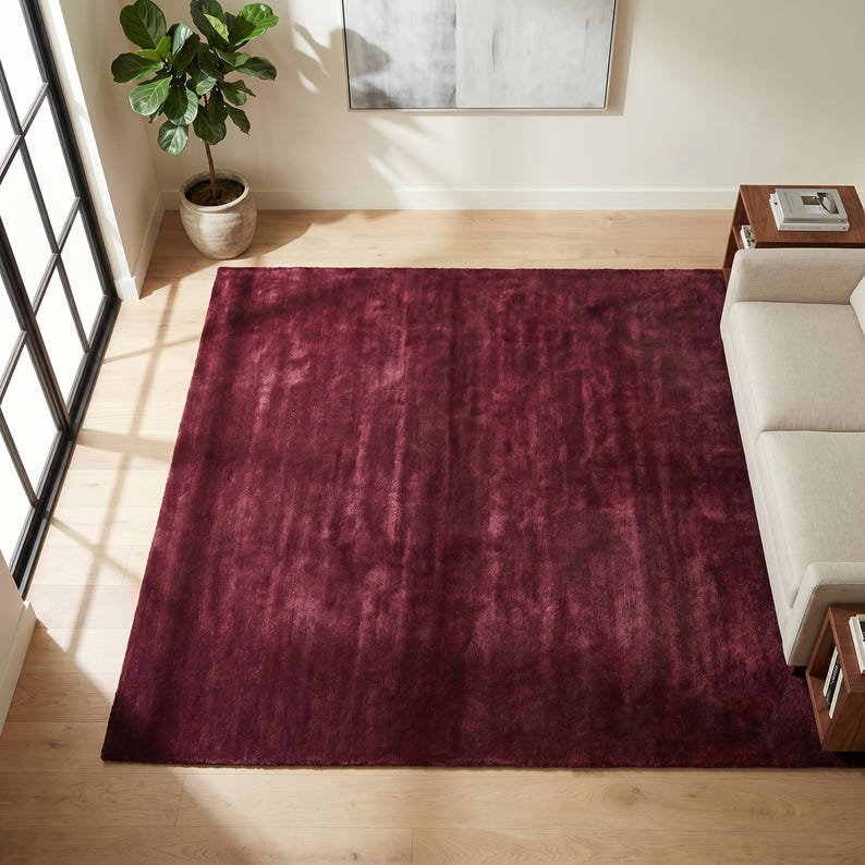 May include: A large, rectangular burgundy rug dominates the room's center. The rug has a soft, textured appearance and is placed on a light-colored hardwood floor. A neutral-toned sofa and side table are partially visible.