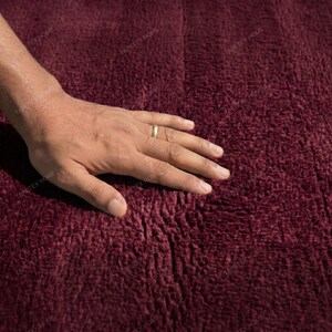 May include: Close-up of a burgundy-colored, textured rug. A hand with a gold ring rests on the rug, showcasing its soft, plush surface. The rug's deep color and texture suggest a cozy, inviting feel, suitable for home decor.