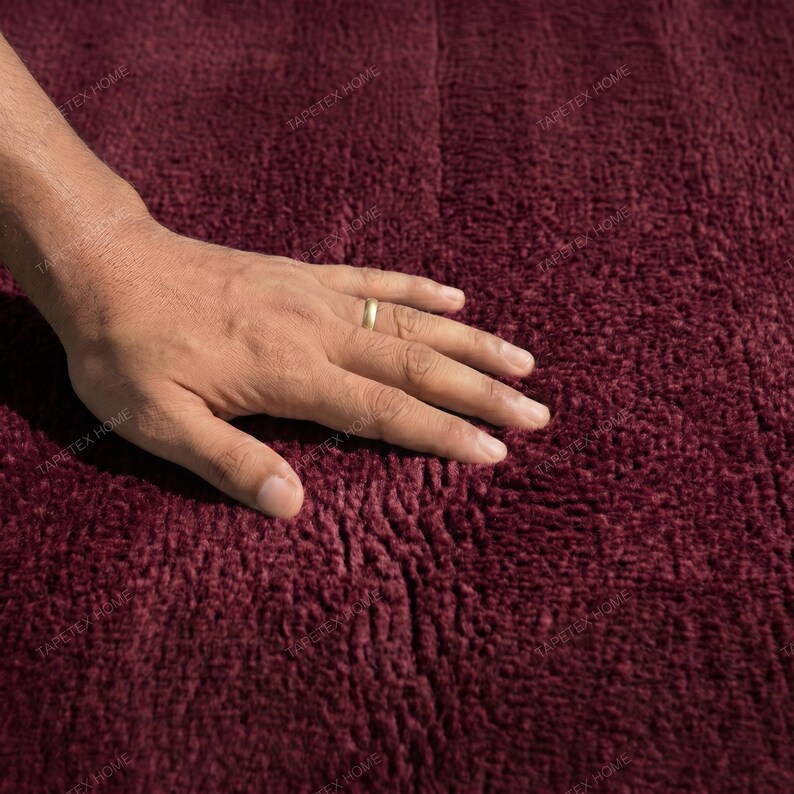May include: Close-up of a burgundy-colored, textured rug. A hand with a gold ring rests on the rug, showcasing its soft, plush surface. The rug's deep color and texture suggest a cozy, inviting feel, suitable for home decor.