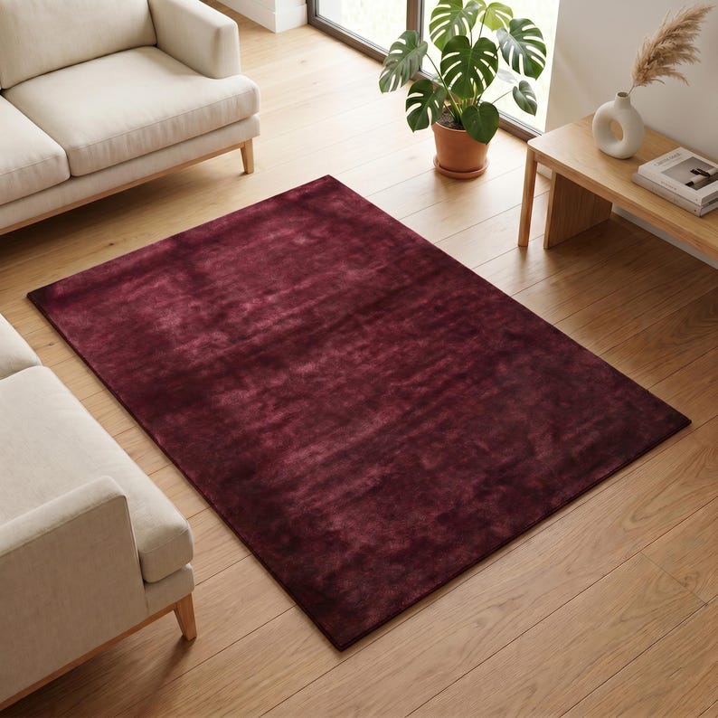 May include: A rectangular, burgundy-colored rug with a textured surface. The rug is placed on a hardwood floor, with a cream-colored sofa visible in the background. The rug adds a touch of warmth and style to the living space.