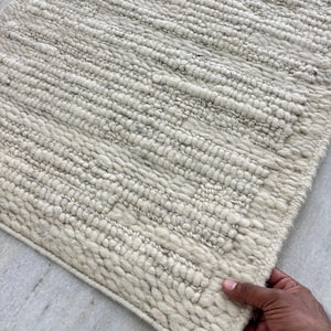 May include: Off-white rectangular rug with a textured, woven design. The rug features a thick pile and a border. The texture appears to be made of wool or a similar material. The rug is held by a hand.