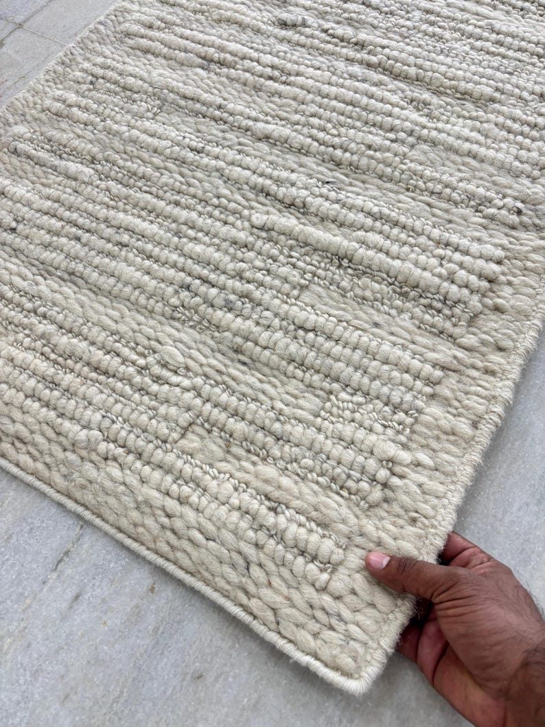 May include: Off-white rectangular rug with a textured, woven design. The rug features a thick pile and a border. The texture appears to be made of wool or a similar material. The rug is held by a hand.