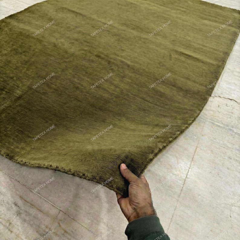 May include: Olive green rug with a textured surface. The rug is rectangular with rounded corners. The rug is being held up by a person's hand. The words "TAPETEX HOME" are printed on the rug.