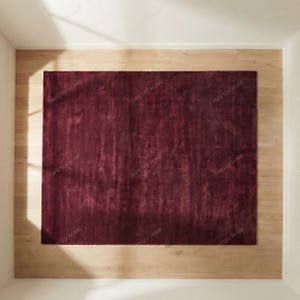 May include: A rectangular, deep burgundy rug with a plush texture. The rug is centered on a light-colored wooden floor, surrounded by white walls. The rug's rich color and soft texture create a warm and inviting atmosphere.