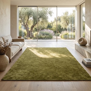 May include: A large, olive green area rug in a modern living room. The rug is rectangular and covers a significant portion of the hardwood floor. A beige sofa and a large window overlooking a garden are also visible.