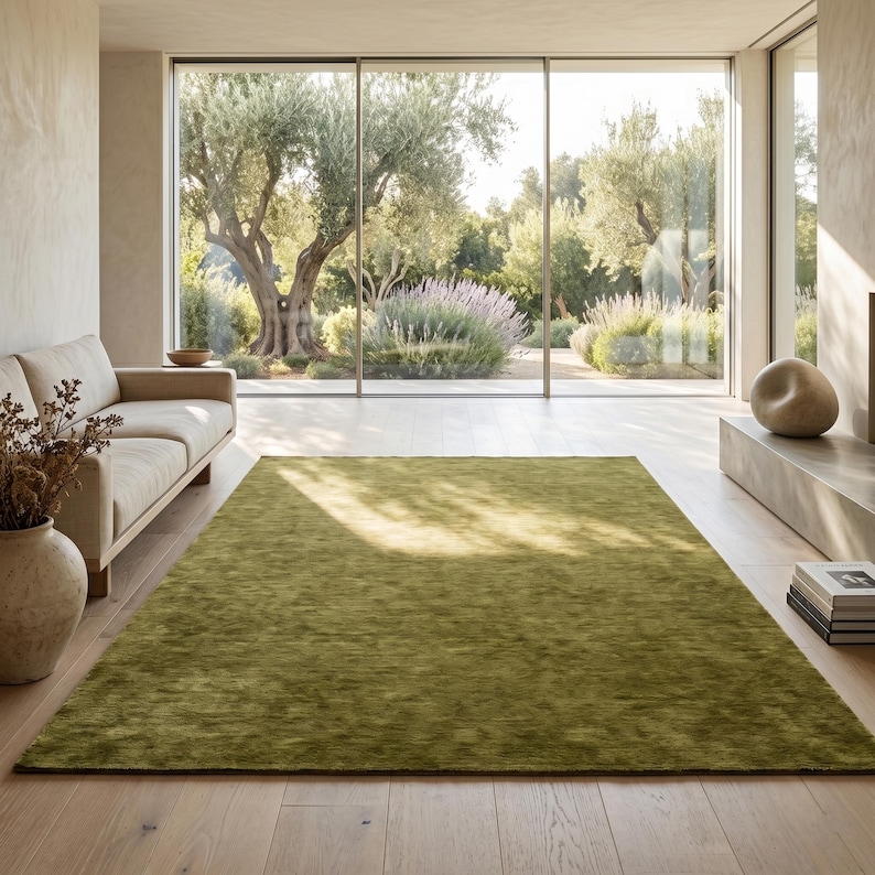 May include: A large, olive green area rug in a modern living room. The rug is rectangular and covers a significant portion of the hardwood floor. A beige sofa and a large window overlooking a garden are also visible.