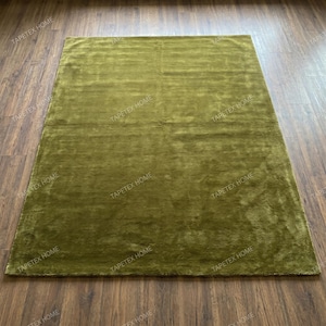 May include: A rectangular olive green rug with a plush, textured surface. The rug is placed on a dark brown wooden floor, creating a contrast in color and texture. The rug appears to be made of a soft material.