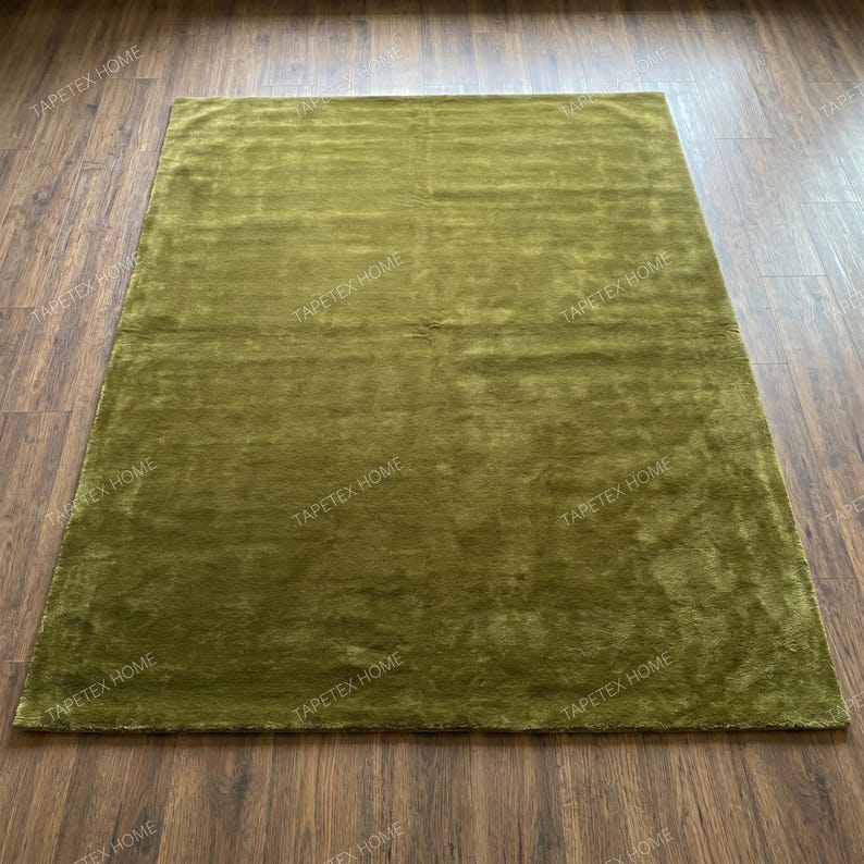 May include: A rectangular olive green rug with a plush, textured surface. The rug is placed on a dark brown wooden floor, creating a contrast in color and texture. The rug appears to be made of a soft material.