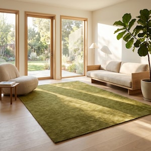 May include: A modern living room with a large, rectangular green rug. The room features a light-colored sofa, a round chair, and a potted plant. Large windows and doors provide natural light and a view of the outdoors.