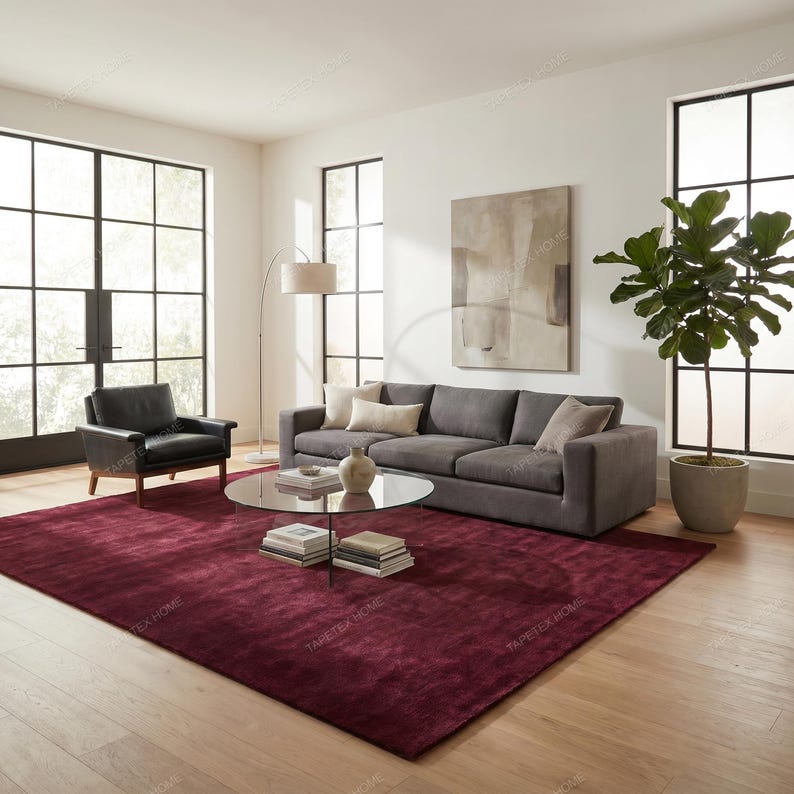 May include: A modern living room featuring a large burgundy area rug, a gray sofa with accent pillows, and a black leather armchair. A glass coffee table displays books and a vase. Large windows and a potted plant complete the scene.