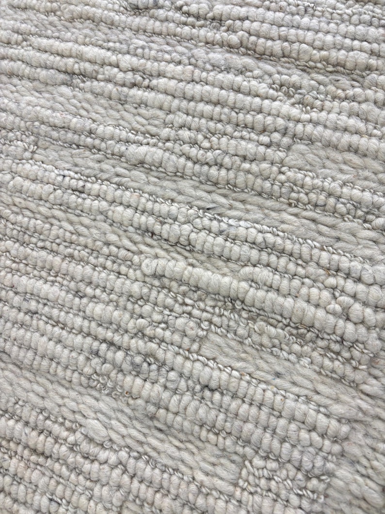 May include: Close-up of a textured, light gray rug. The rug has a ribbed pattern with a soft, plush appearance. The fibers are tightly woven, creating a durable and comfortable surface. The neutral color makes it suitable for various interior design styles.
