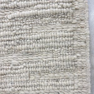 May include: Close-up of a textured, off-white rug. The rug features a repeating pattern of raised, horizontal lines and a geometric design. The material appears to be a thick, woven fabric, creating a cozy and inviting aesthetic.