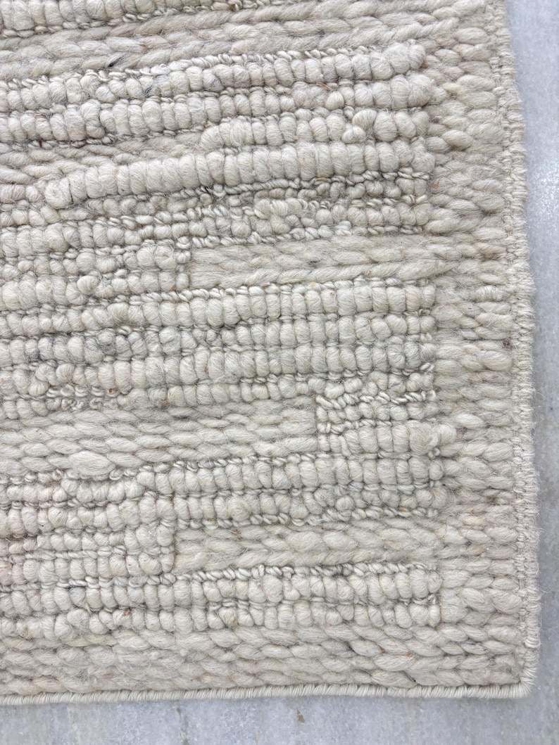 May include: Close-up of a textured, off-white rug. The rug features a repeating pattern of raised, horizontal lines and a geometric design. The material appears to be a thick, woven fabric, creating a cozy and inviting aesthetic.