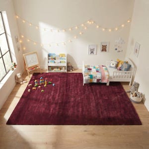 May include: A burgundy rectangular area rug in a child's bedroom. The room features a white bed with a colorful quilt, a white bookshelf, and a wooden easel with toys scattered on the floor. String lights hang from the ceiling.