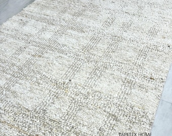 Pode incluir: Um tapete retangular branco sujo com uma superfície tecida e texturizada. O tapete apresenta um padrão quadriculado em um tom mais claro, criando um contraste sutil. As palavras "TAPETEX HOME" são impressas em preto nos cantos superior esquerdo e inferior direito.
