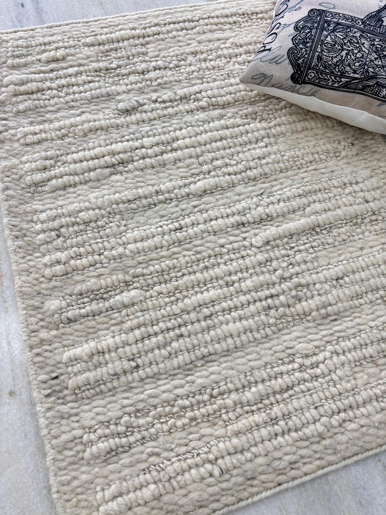 May include: A rectangular, off-white rug with a textured, looped pile. The rug has a horizontal striped pattern and a soft, neutral color. A decorative pillow with a black and white design rests on the rug.
