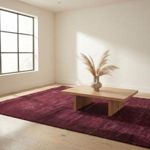 May include: A modern living room featuring a large, rectangular, burgundy rug. A light wood coffee table sits on the rug, with a vase of dried plants on top. A large window is visible in the background.