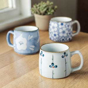 May include: Three ceramic mugs with floral patterns. One is light blue with a white flower, another has a blue and white design, and the third is white with blue flowers and red accents. All have brown rims.