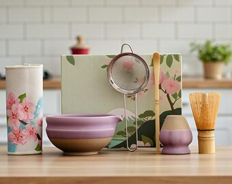 May include: A matcha tea set with a lavender ceramic bowl, a bamboo whisk, a stainless steel strainer, and a bamboo scoop. The set includes a container with a floral design and a lavender and tan ceramic tea cup holder. The background is a kitchen setting.