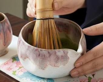 May include: A ceramic matcha bowl with a white and purple glaze, a bamboo whisk, and a small tea strainer. The bowl is filled with green matcha tea. The whisk is being used to mix the tea. The scene is set on a floral patterned mat.