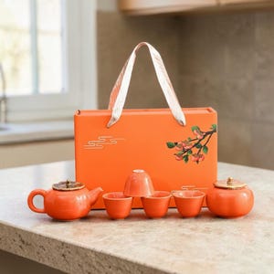 Könnte beinhalten: Ein orangefarbenes Teeservice mit Teekanne, Tassen und Zuckerdose, präsentiert in einer passenden Geschenkbox mit Griff. Die Box ist mit einem floralen Zweigdesign verziert. Das Teeservice steht auf einer hellen Arbeitsplatte.