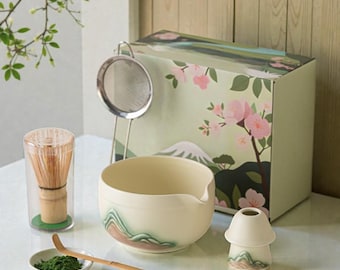 May include: A matcha tea set with a bamboo whisk, ceramic bowl, sifter, scoop, and a small ceramic holder. The set includes a box with a floral and mountain design. The bowl and holder have a mountain design. The matcha powder is green.