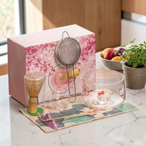 Può includere: Una scatola rosa con un design di fiori di ciliegio contiene un set da tè matcha. Il set include una frusta di bambù, una caraffa di vetro, due bicchieri piccoli, un colino di metallo e un tappetino floreale. I bicchieri presentano un design di Hello Kitty.