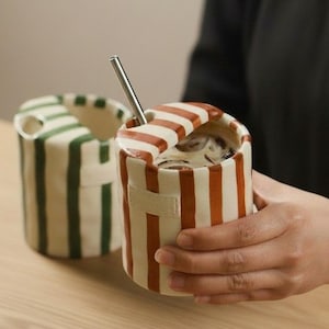 Puede incluir: Una taza de cerámica a rayas con tapa y pajita de metal. La taza tiene una base crema con rayas verticales naranjas. Se ve otra taza con rayas verdes. La taza la sostiene una persona.
