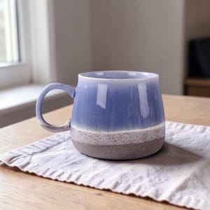 May include: A handmade ceramic mug with a blue glazed top, a white speckled middle band, and a gray base. The mug has a rounded shape and a curved handle. It sits on a white cloth on a wooden table.
