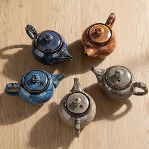 Puede incluir: Cinco teteras de cerámica en tonos azules, marrones y grises dispuestas sobre una superficie de madera. Cada tetera tiene un cuerpo redondeado, un pico, un asa y una tapa. Las teteras tienen un patrón moteado.