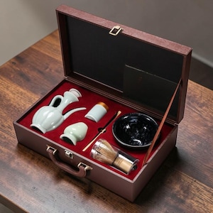 May include: A brown, open carrying case with a red velvet interior holds a tea ceremony set. The set includes a light green ceramic pitcher, a small bowl, a whisk, and other tea preparation tools. The case is on a wooden surface.