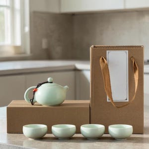 Puede incluir: Un juego de té de cerámica verde claro con una tetera, cuatro tazas pequeñas y una caja de regalo. La tetera tiene un asa negra y un adorno rojo. La caja de regalo es marrón con una etiqueta blanca y un asa beige.