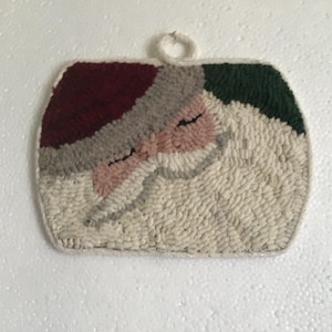 May include: A rectangular punch needle wall hanging featuring a stylized Santa Claus face. The design includes a deep red hat, a light gray brim, a peach-colored face with closed eyes, and a large white beard. A small white loop is attached at the top.