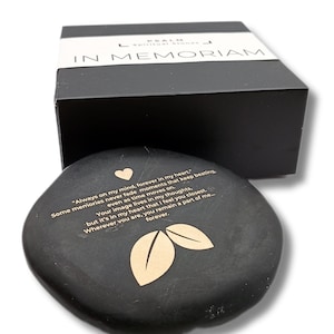 Op de afbeelding: Een gladde, platte, zwarte steen met goudkleurige tekst die luidt: "Always on my mind, forever in my heart." Het bevat een klein gouden hartsymbool en twee gouden bladsymbolen. De steen ligt voor een zwarte doos met het opschrift "IN MEMORIAM."